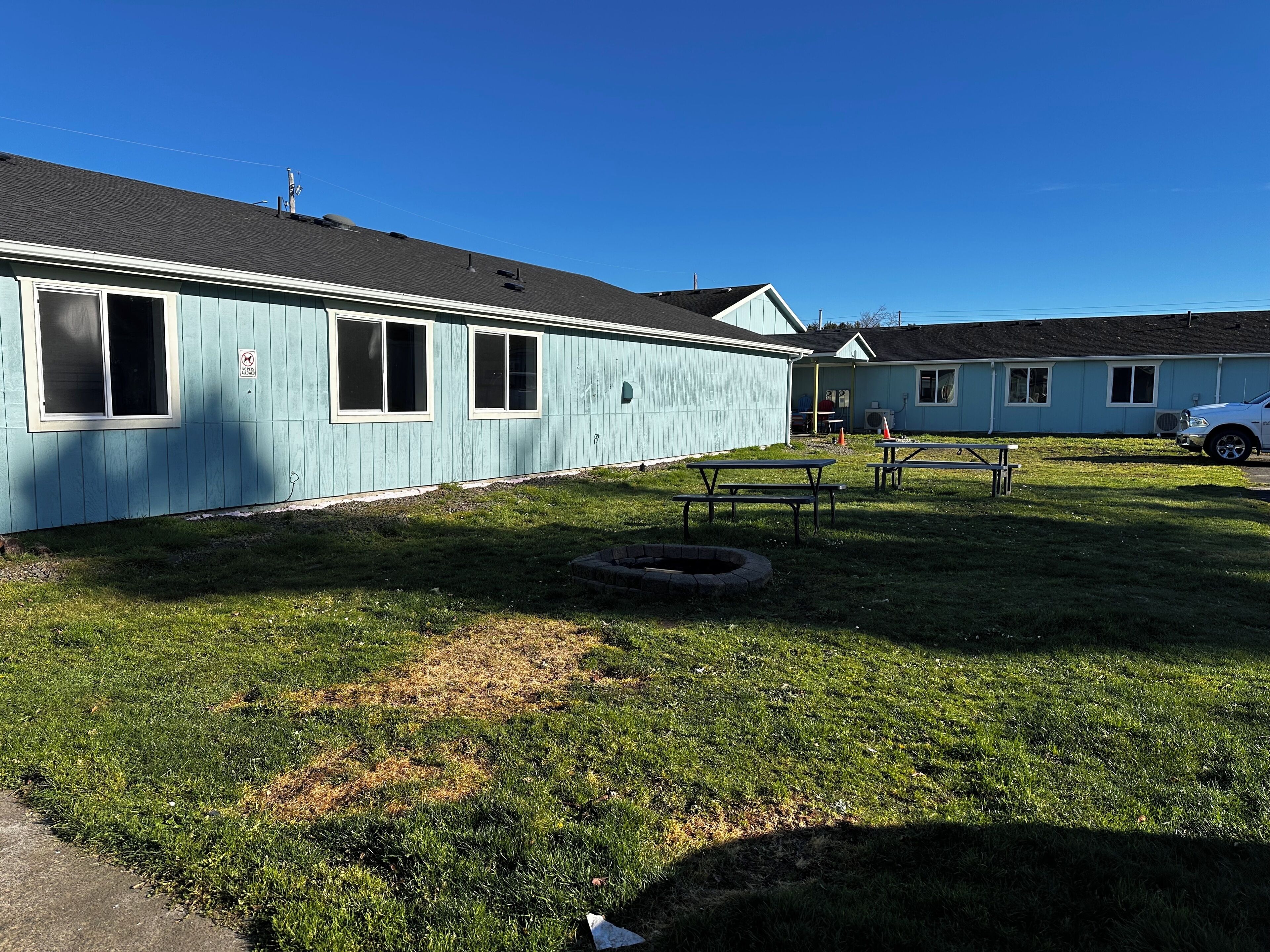 A light blue building with a flat roof, a grassy yard, picnic tables, and a white car parked nearby.
