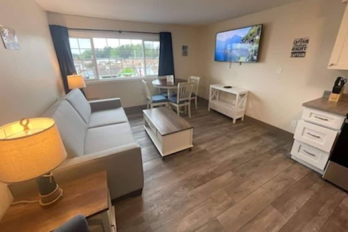 Una sala de estar moderna con sofá, mesa de centro y televisor. Se ve una ventana con cortinas, una mesita y un televisor en la pared.