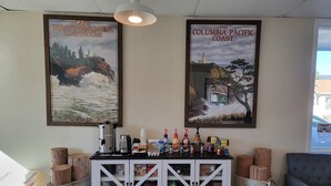 A reception area with a white cabinet displaying various items, two framed posters of coastal scenes, and a hanging light fixture. - The Coastal Inn and Suites (Long Beach)