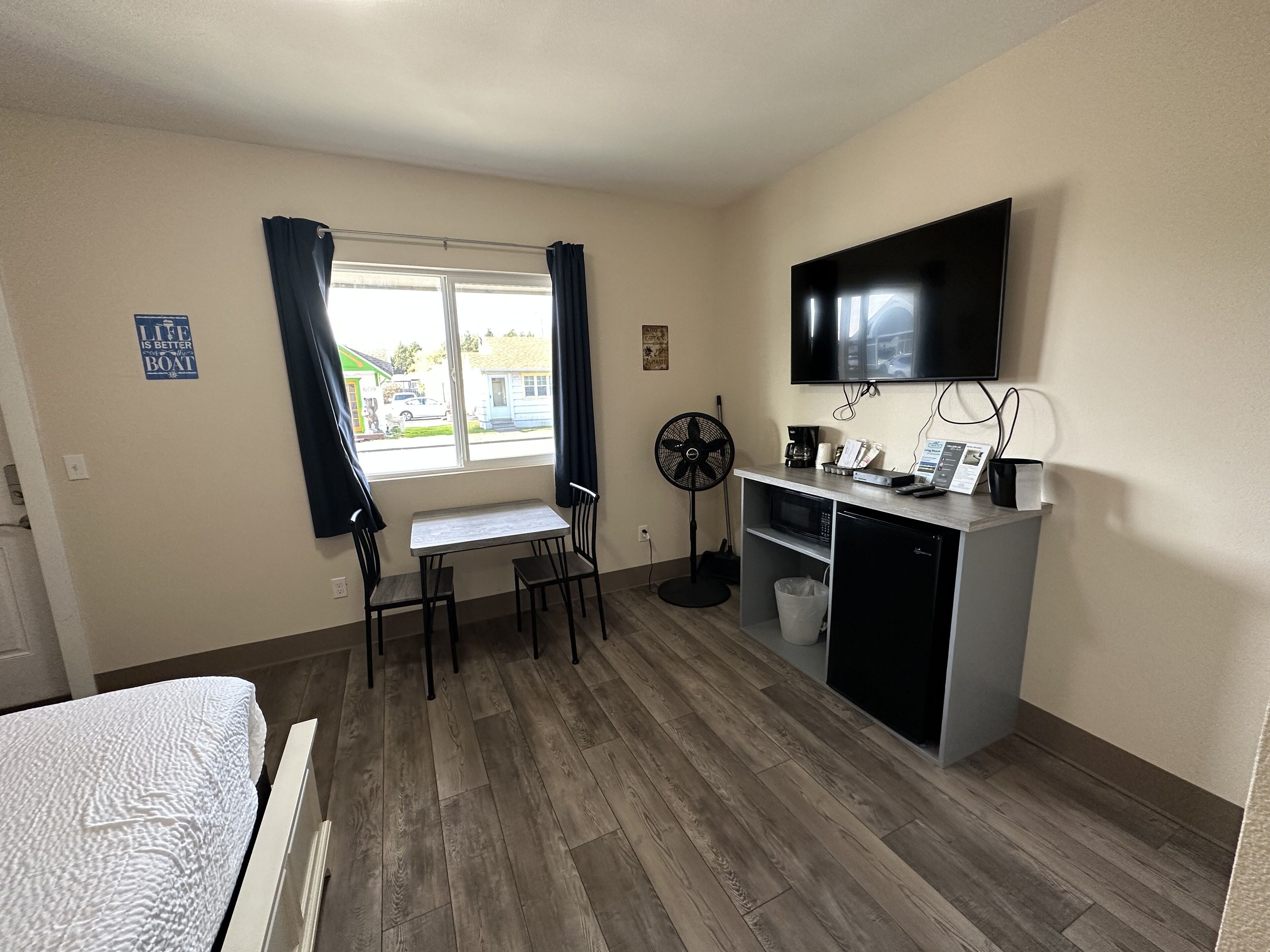 Une chambre d’hôtel compacte comprenant un lit, une petite table avec deux chaises, un bureau avec une télévision, un four à micro-ondes et un ventilateur.