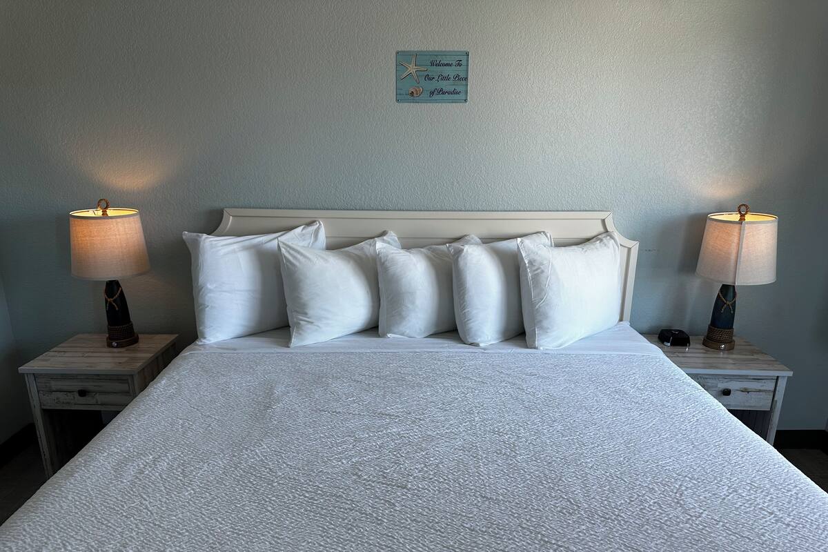 Una cama bien hecha con sábanas blancas y varios almohadones, flanqueada por dos mesitas de noche con lámparas y un pequeño cajón.