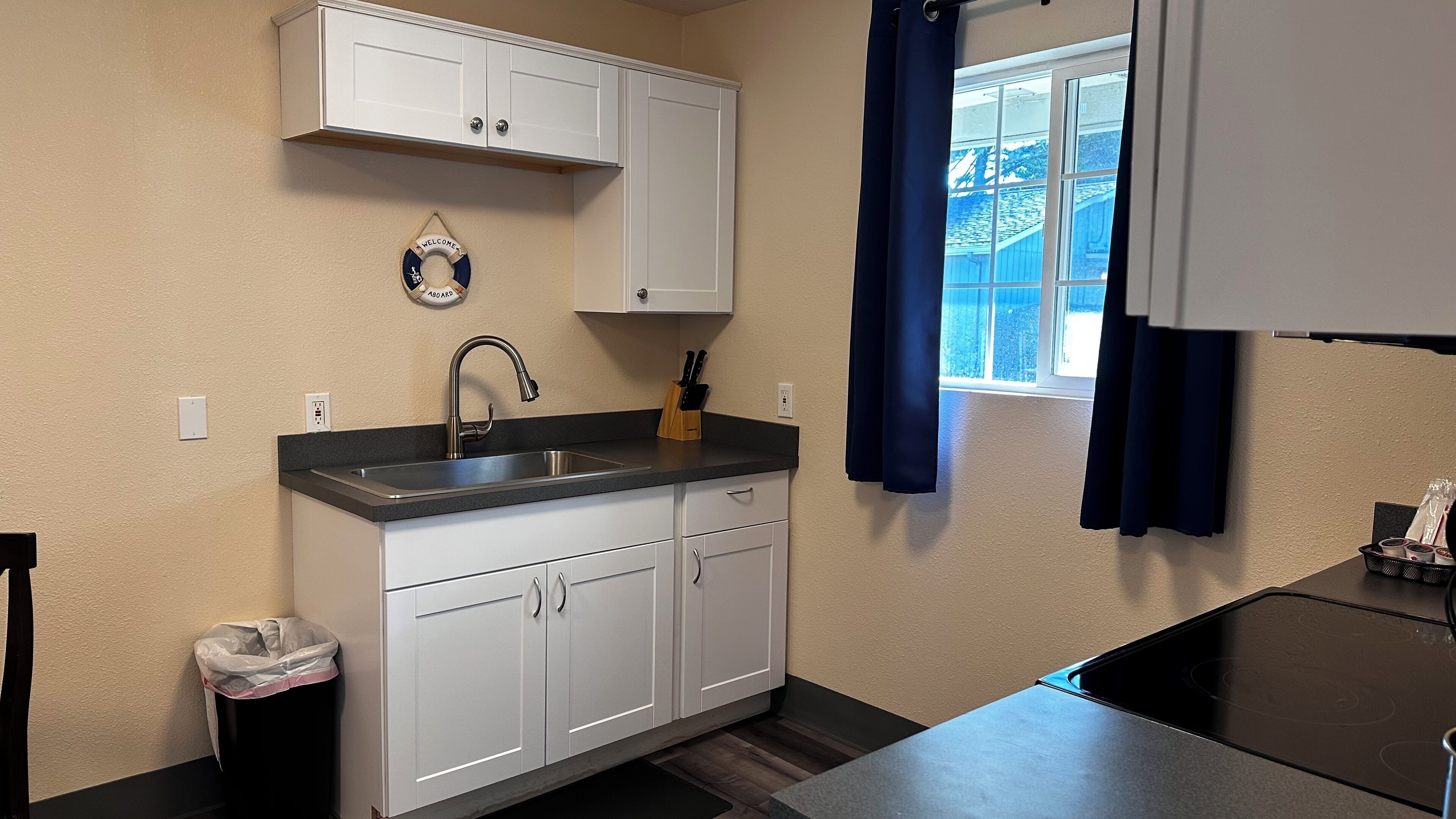 Una cocina con armarios blancos, un lavamanos de acero inoxidable, un ventanal con cortinas azules y vistas al exterior.