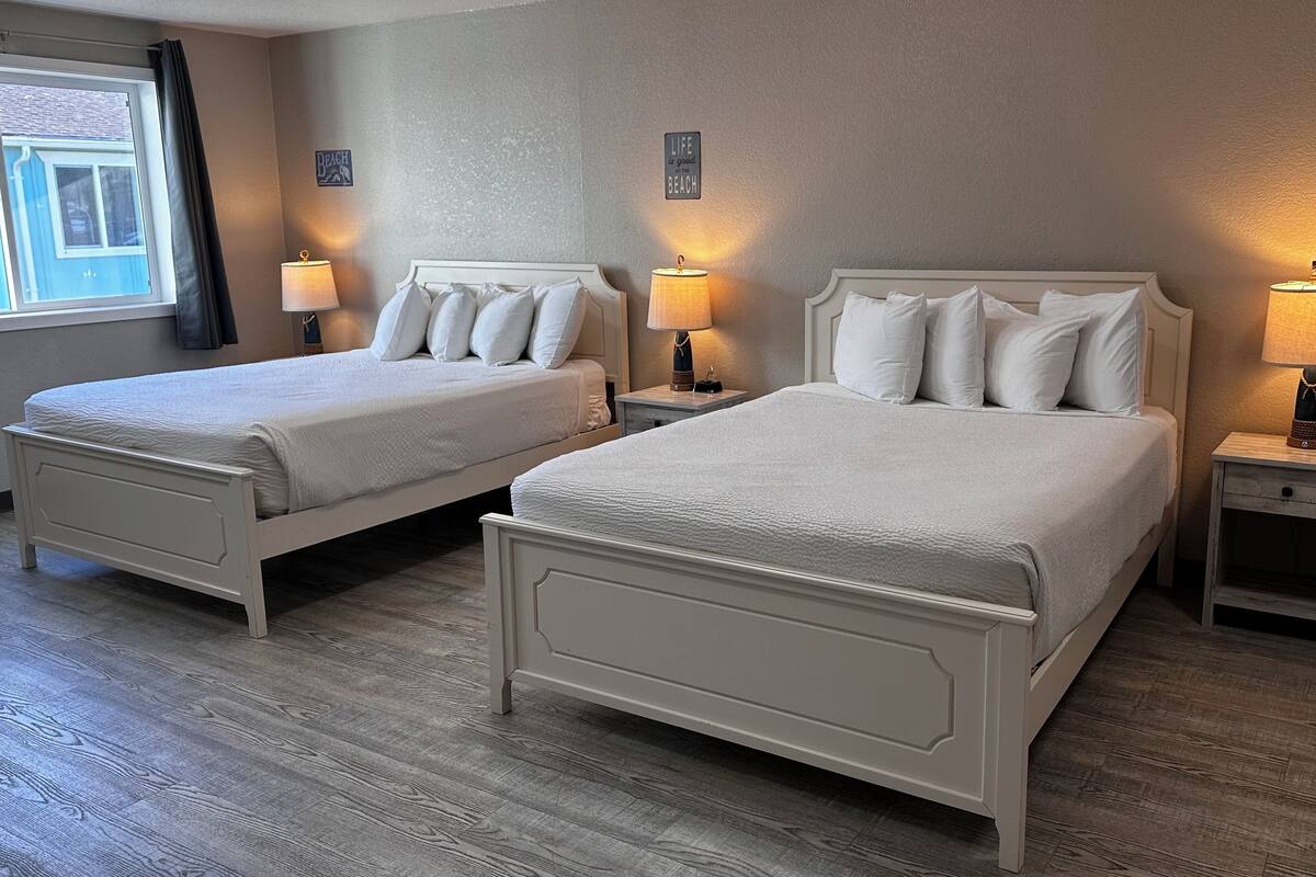 Una habitación de hotel con dos camas individuales, cada una con una mesita de noche y lámpara, una ventana con cortinas y una pared decorada con un cartel con motivos playeros.
