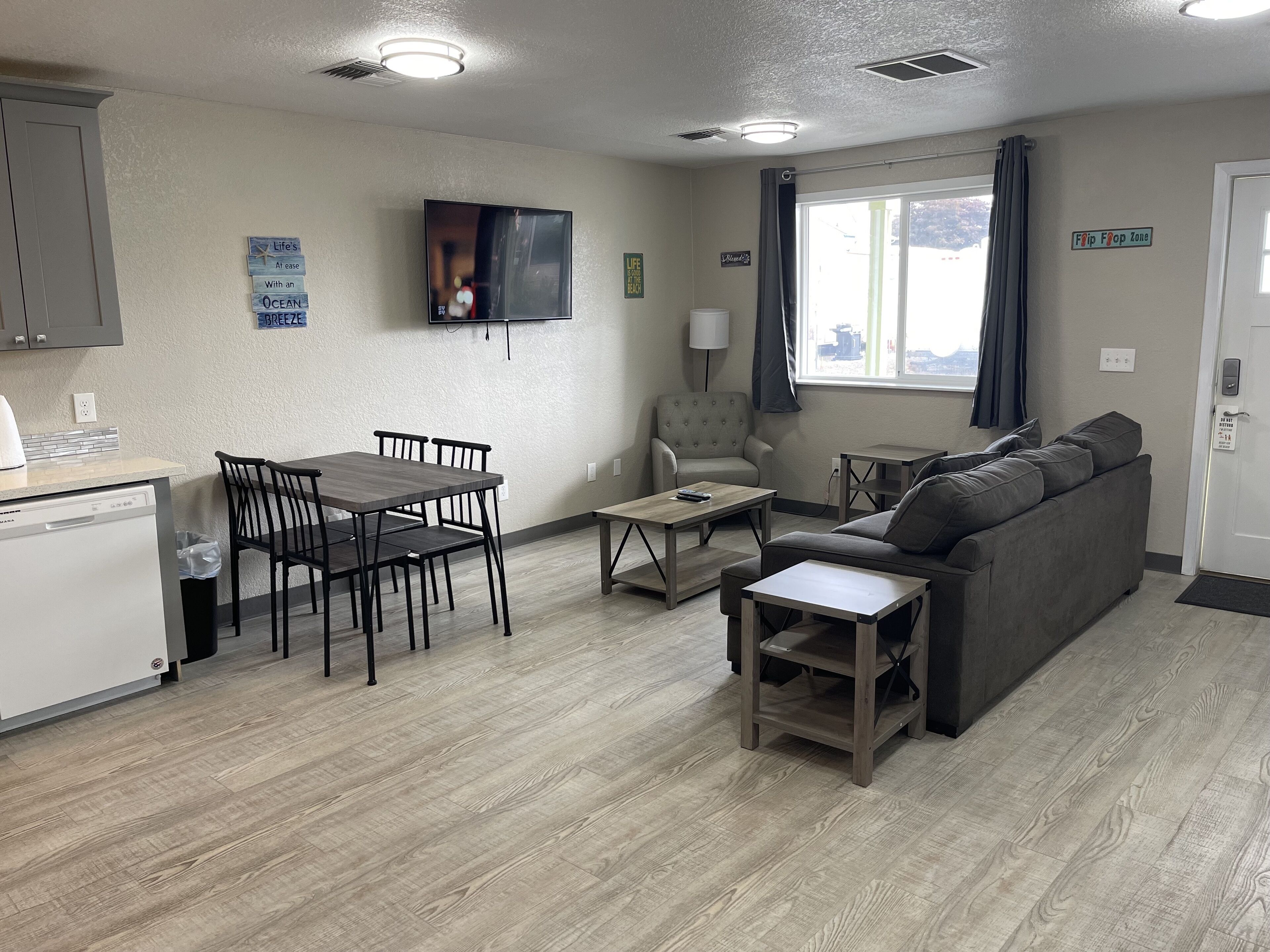 A modern living room with a sofa, coffee table, and dining table. There is a television mounted on the wall, a window with curtains, and a door with a sign.