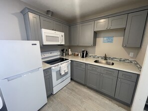 A compact kitchen with grey cabinets, a white refrigerator, a microwave, a stove, and a sink. - The Coastal Inn and Suites (Long Beach)