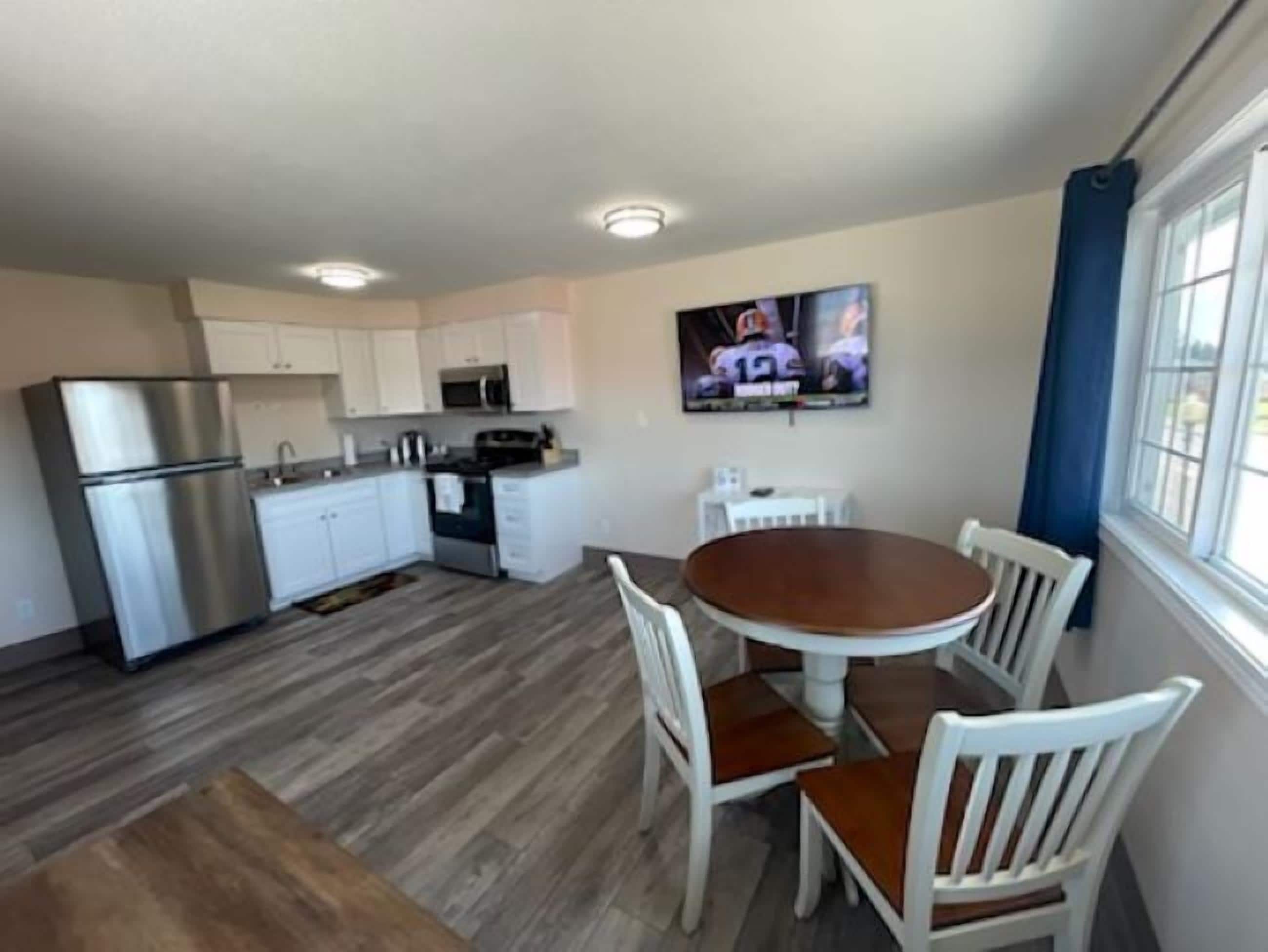 A compact kitchen with white cabinets, a stainless steel refrigerator, and a microwave. There's a dining area with a round wooden table and white chairs. A TV is mounted on the wall above the table.