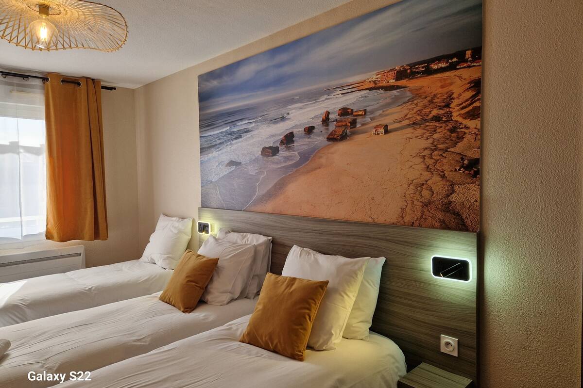 A hotel room with two beds, a beach mural, and a modern pendant light.