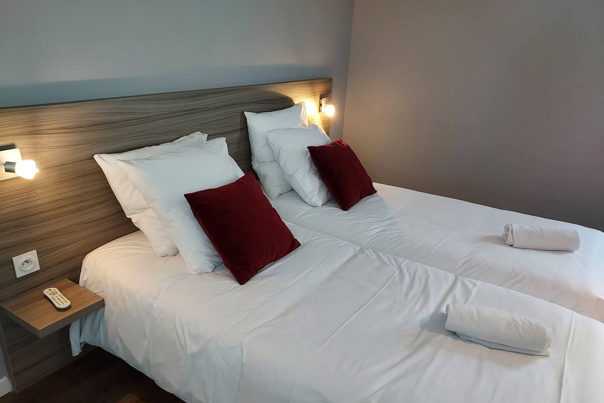 A hotel room with two beds, white bedding, red pillows, a wooden headboard, bedside lamps, a remote control, and a folded towel on the right bed.