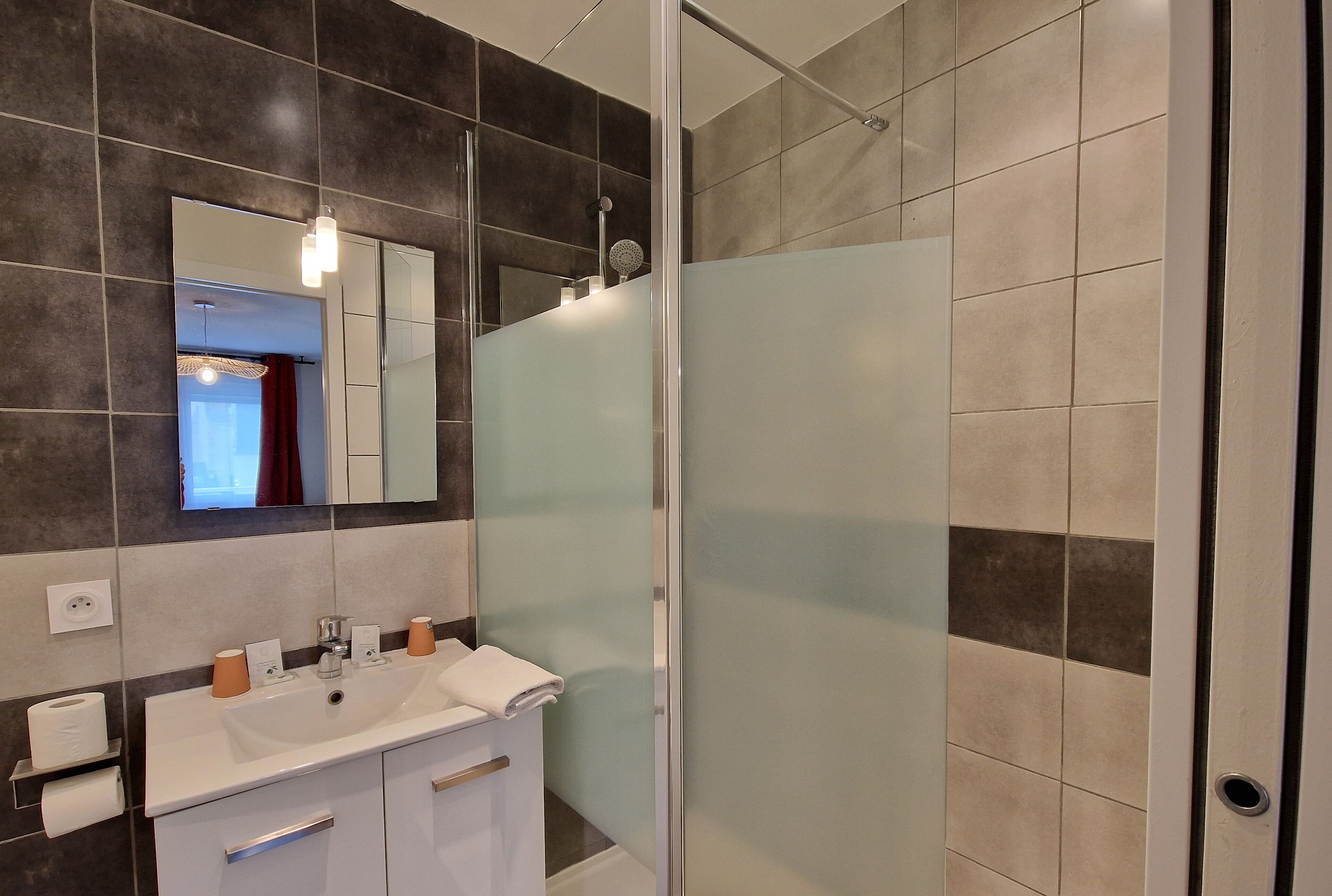 Un baño moderno con cabina de ducha de vidrio, un lavabo blanco con espejo y paredes cubiertas de azulejos oscuros.