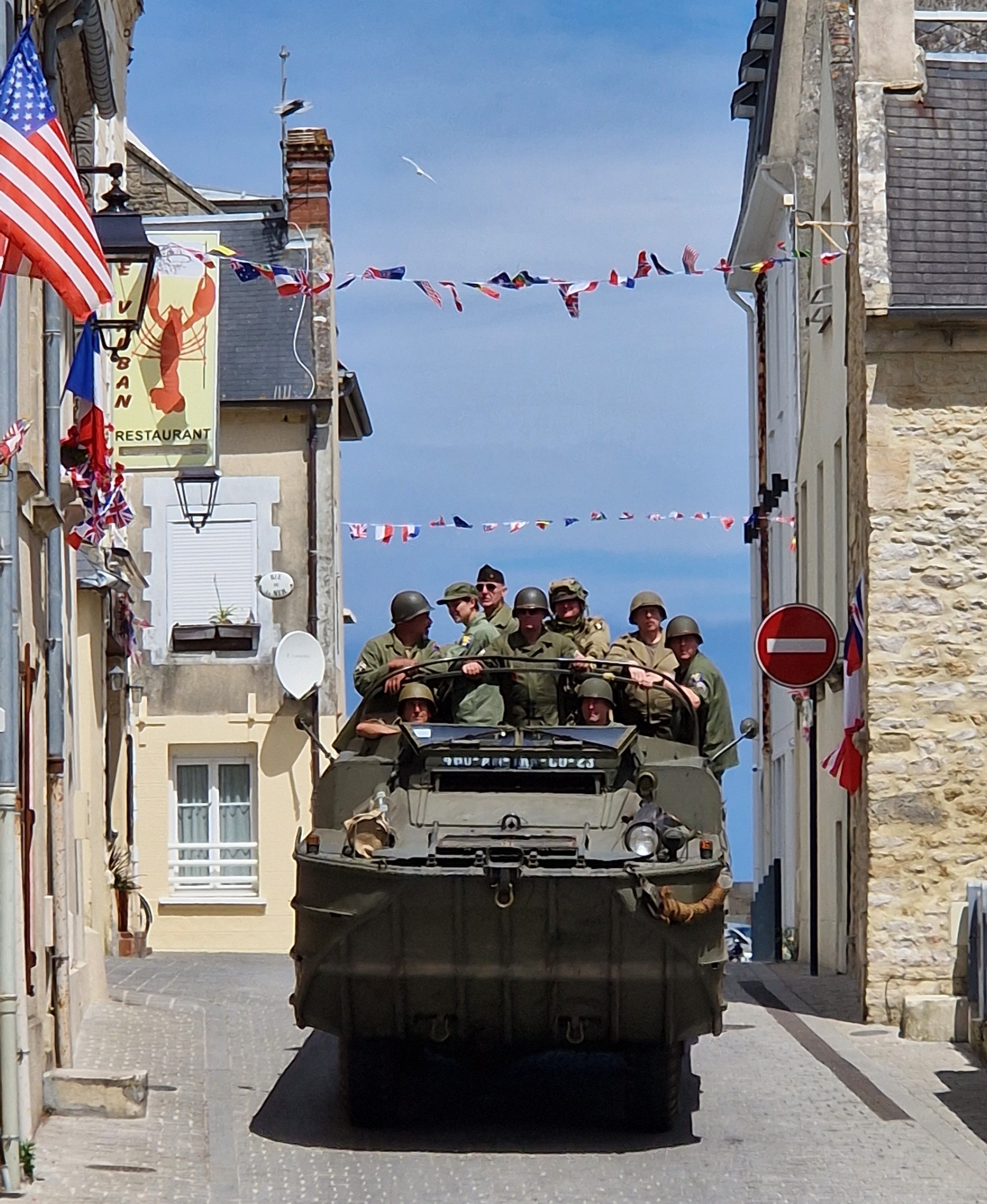 A military vehicle with soldiers in a narrow street, surrounded by buildings with flags and a 'RESTAURANT' sign.