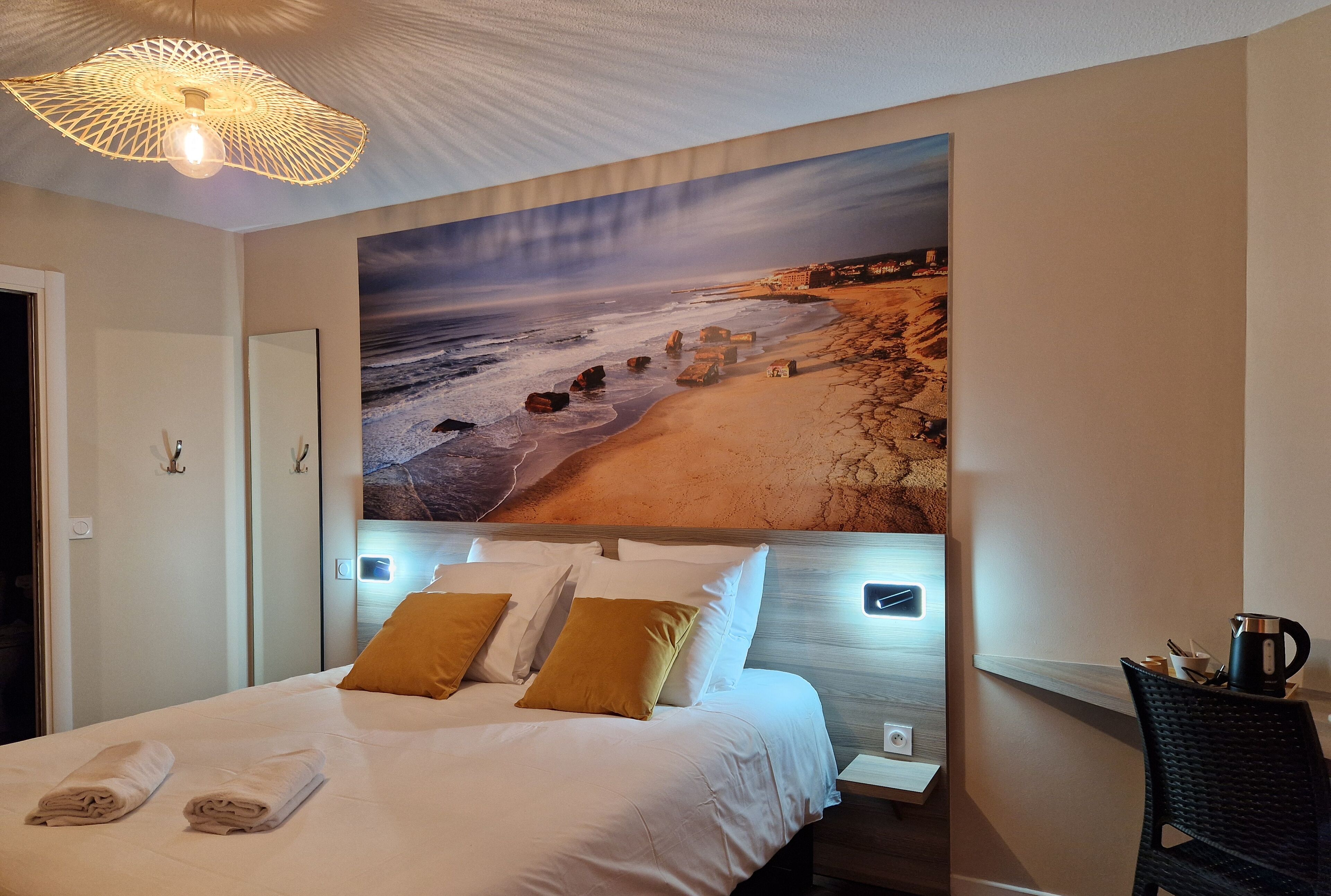 Ein Hotelzimmer mit einer großen Strandmalerei an der Wand, einem ordentlich bezogenen Bett mit weißer Bettwäsche und einer modernen Lampe darüber.