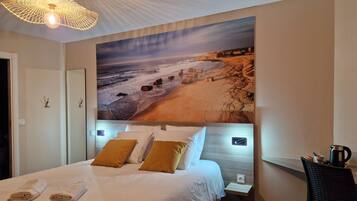 Ein Hotelzimmer mit einer großen Strandmalerei an der Wand, einem ordentlich bezogenen Bett mit weißer Bettwäsche und einer modernen Lampe darüber.