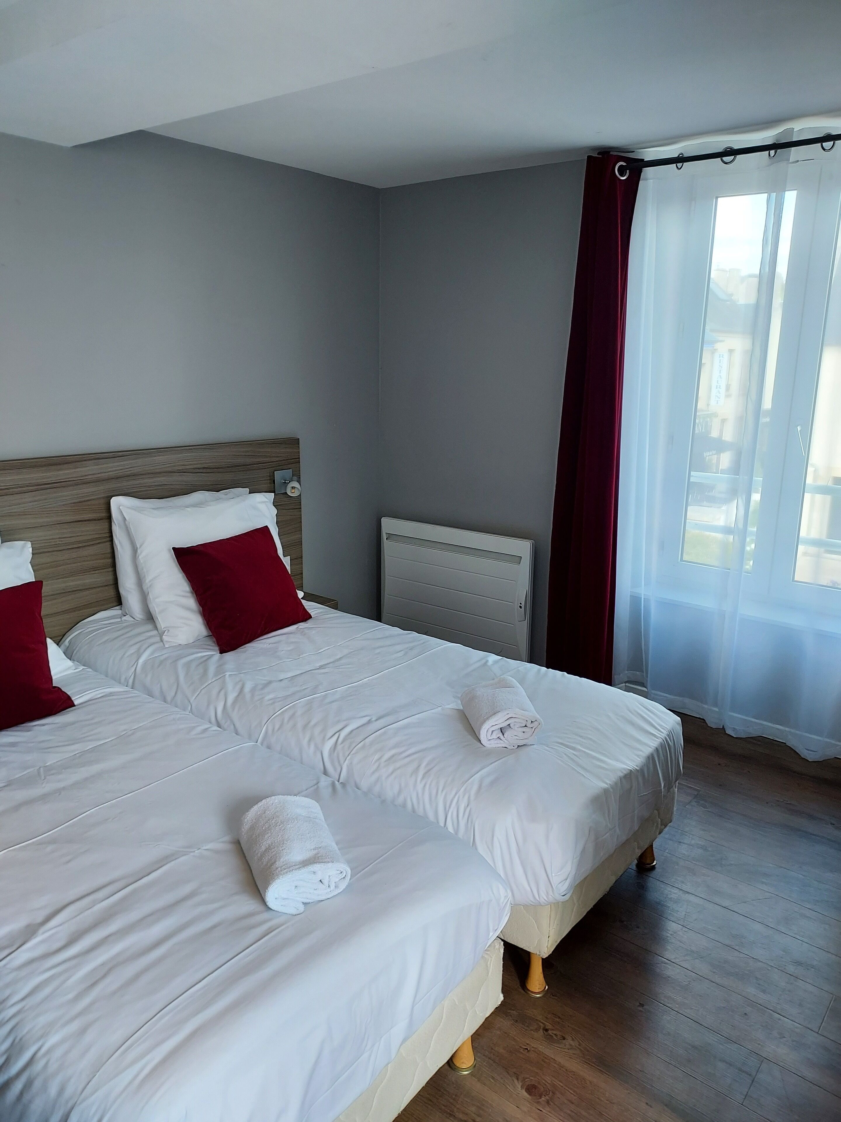 A hotel room with two beds, a wooden headboard, a radiator, and a window with sheer curtains.