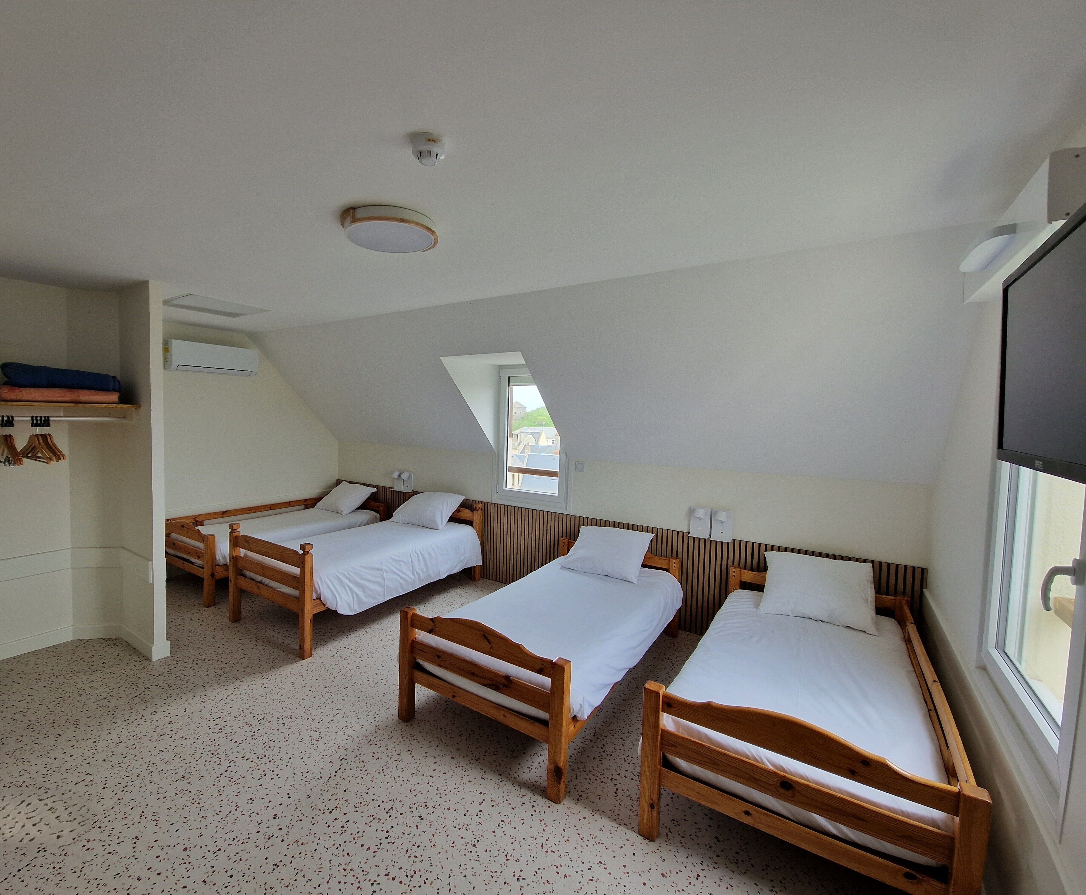 Une chambre de dortoir avec trois lits en bois, un ventilateur de plafond, un appareil de climatisation et une fenêtre laissant entrer la lumière naturelle.