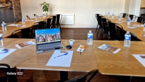 Un salón de conferencias con una mesa de madera, sillas negras, una computadora portátil, botellas de agua y una planta.