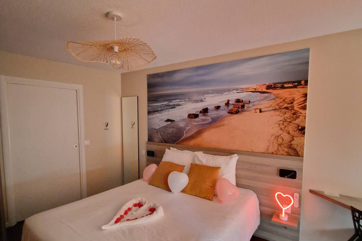 A hotel room with a bed, a beach mural, a ceiling fan, and a bedside table with a neon heart sign.