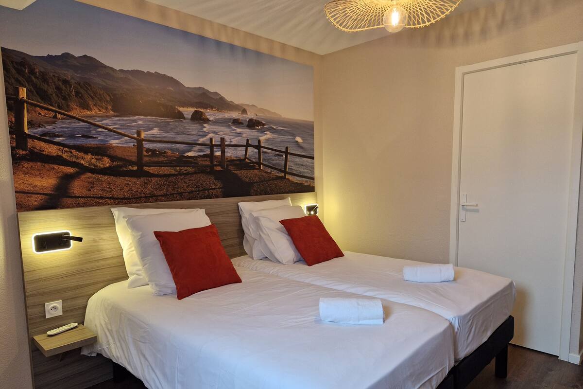 A double bed with white linens and red pillows, a nightstand with a remote control, and a wall-mounted light fixture.