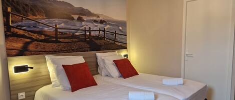 A double bed with white linens and red pillows, a nightstand with a remote control, and a wall-mounted light fixture.