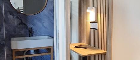 A modern bathroom with a marble wall, a round mirror, a sink, a wooden vanity, a television mounted above the mirror, and a wall-mounted light fixture.