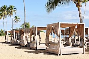 A row of beige canopy beds with curtains, arranged on a sandy beach with palm trees in the background. - Royalton CHIC Punta Cana, An Autograph Collection All-Inclusive Resort & Casino – Adults Only (Punta Cana)