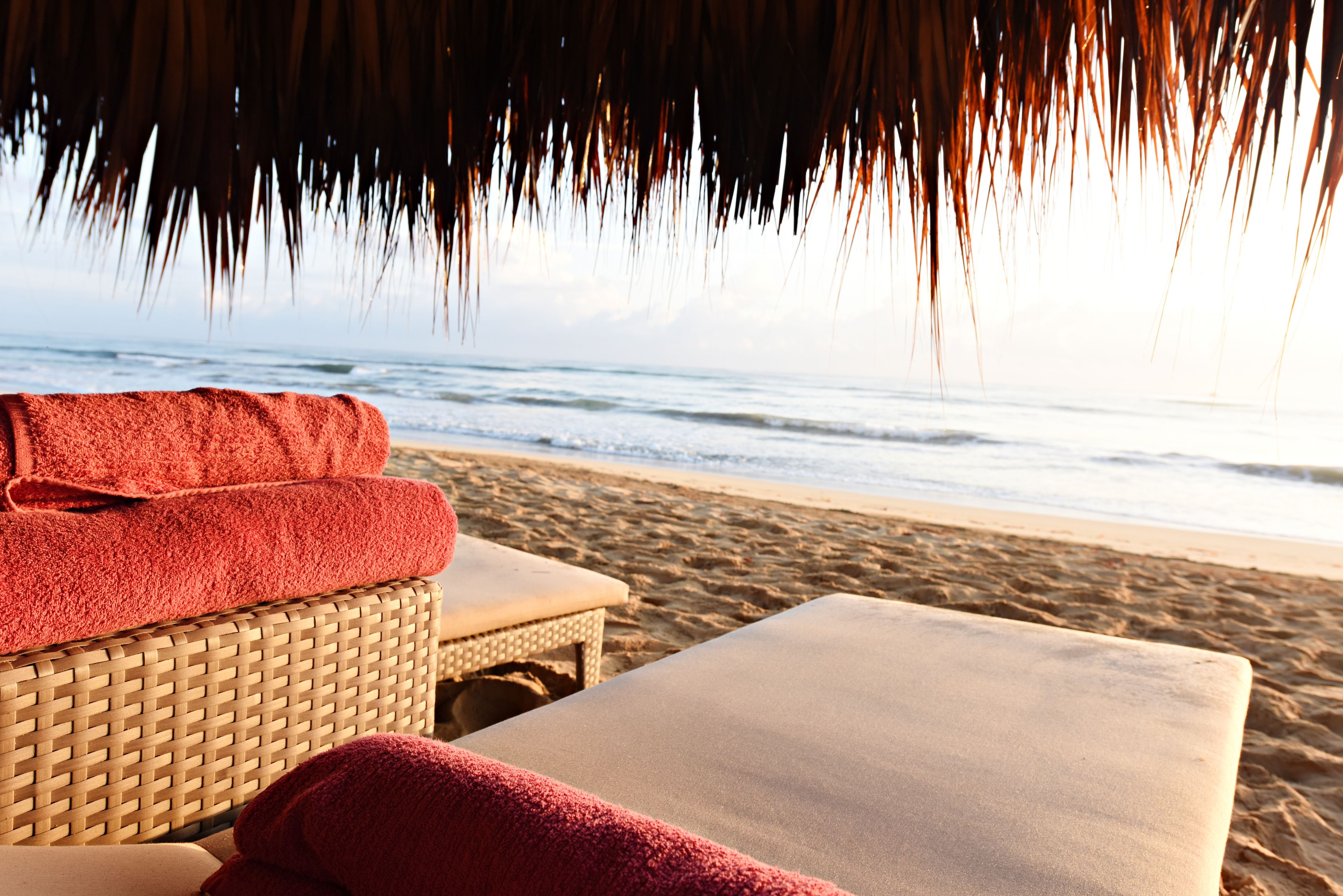 Un salon de plage avec un fauteuil en osier et un banc rembourré, une serviette rouge et une vue sur l’océan.