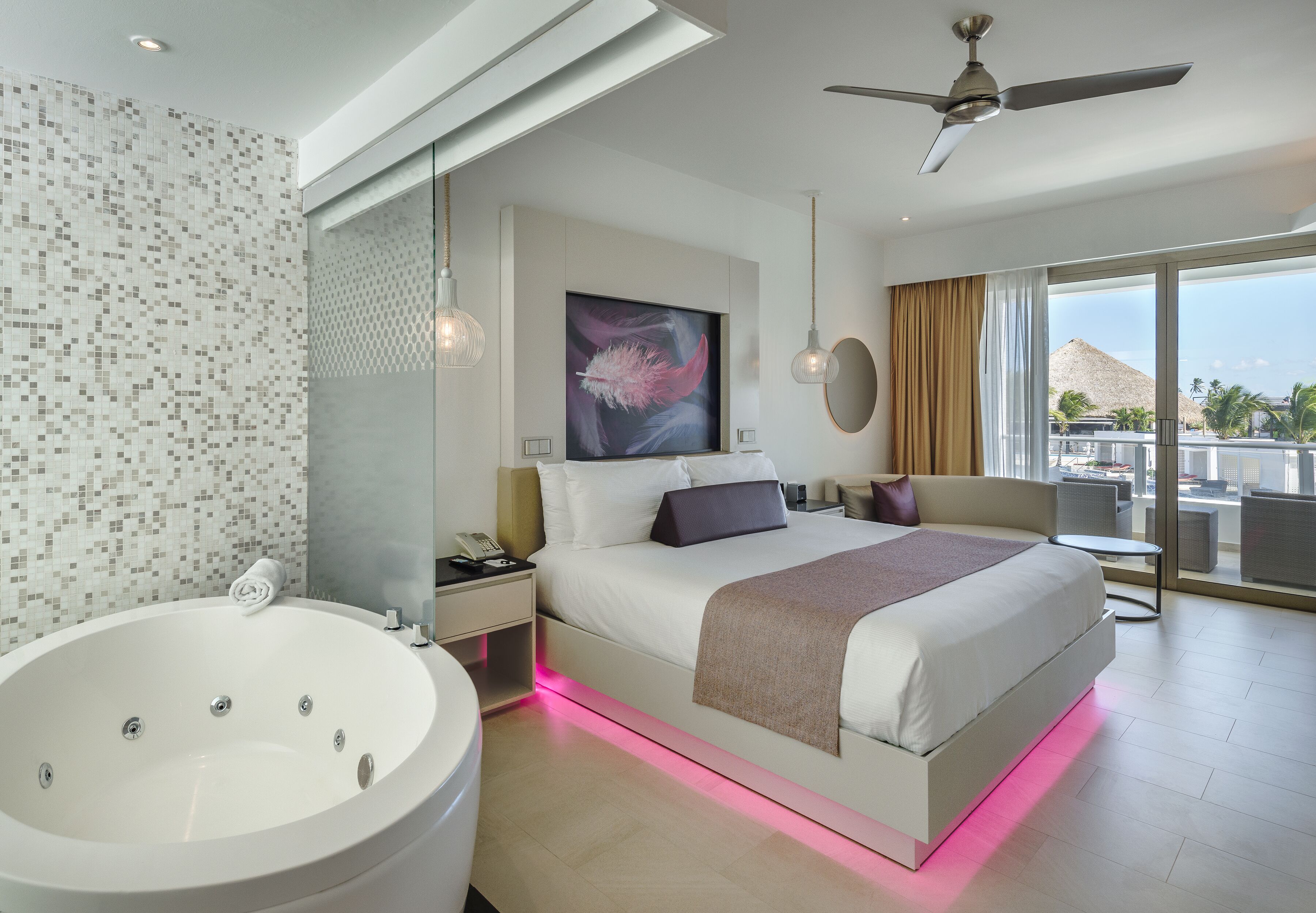 Ein modernes Hotelzimmer mit einem großen Bett, einer Badewanne und einem Balkon mit Blick auf Palmen und ein Gebirge.