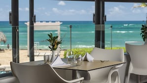 A dining area with a table set for two, overlooking a beach with the ocean and a thatched hut in the background.