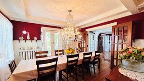 A dining room with a wooden table, chairs, a chandelier, and a view of the kitchen and garden. - Chestnut Inn Bed and Breakfast (Niagara Falls)