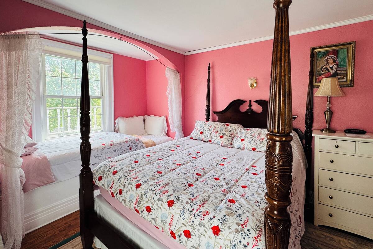 A four-poster bed with floral bedding, a nightstand with a lamp, and a window with a view of greenery.