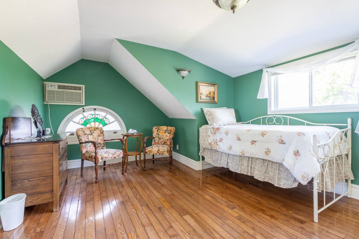 A room with a sloped ceiling, a bed with floral bedding, two floral armchairs, a small table, and a window with a view of greenery.