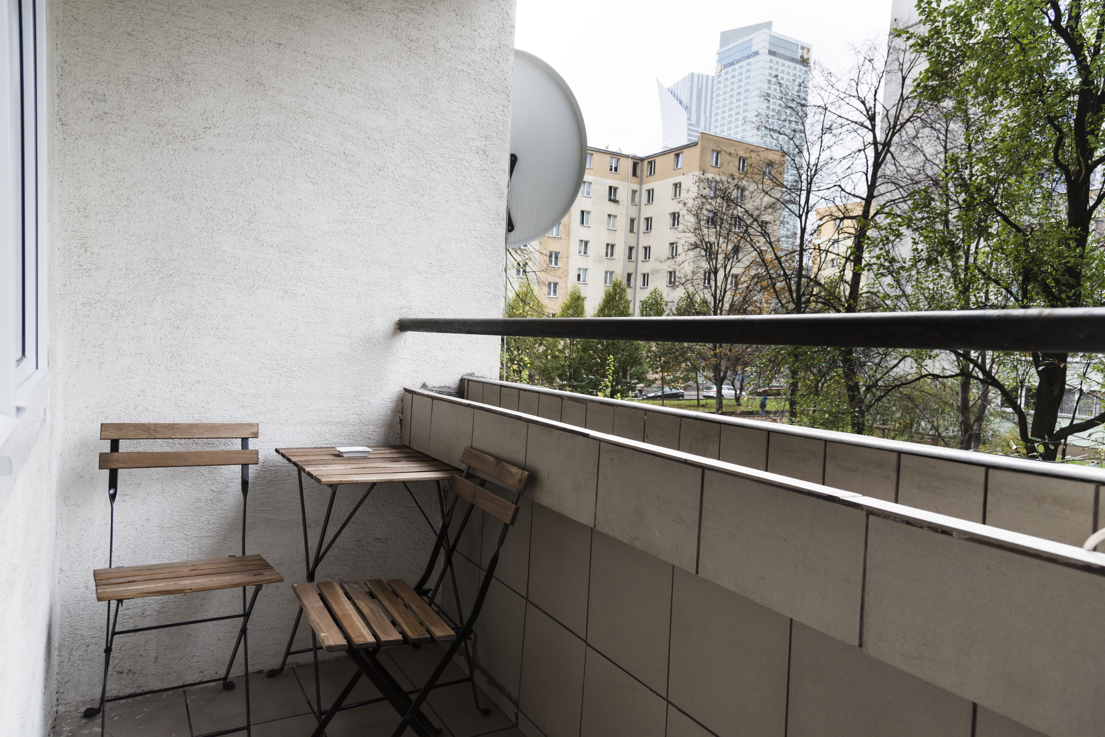 Un balcon avec deux tables et des chaises en bois, une antenne parabolique et une vue sur des immeubles et des arbres.