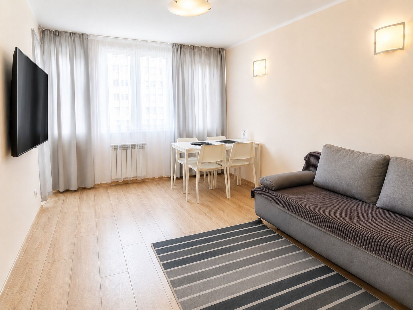 A modern living room with a flat-screen TV, a sofa, a dining table with chairs, and a window with curtains.