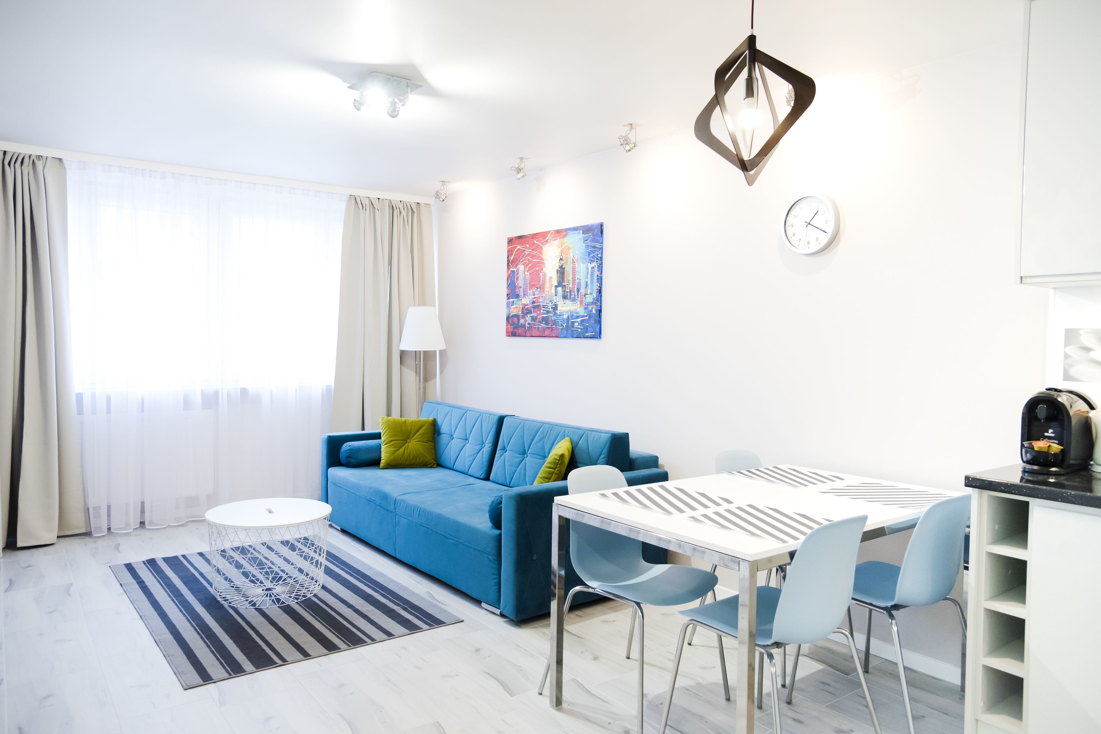 A modern living room with a blue sofa, a dining table with chairs, a coffee machine, and a painting on the wall.