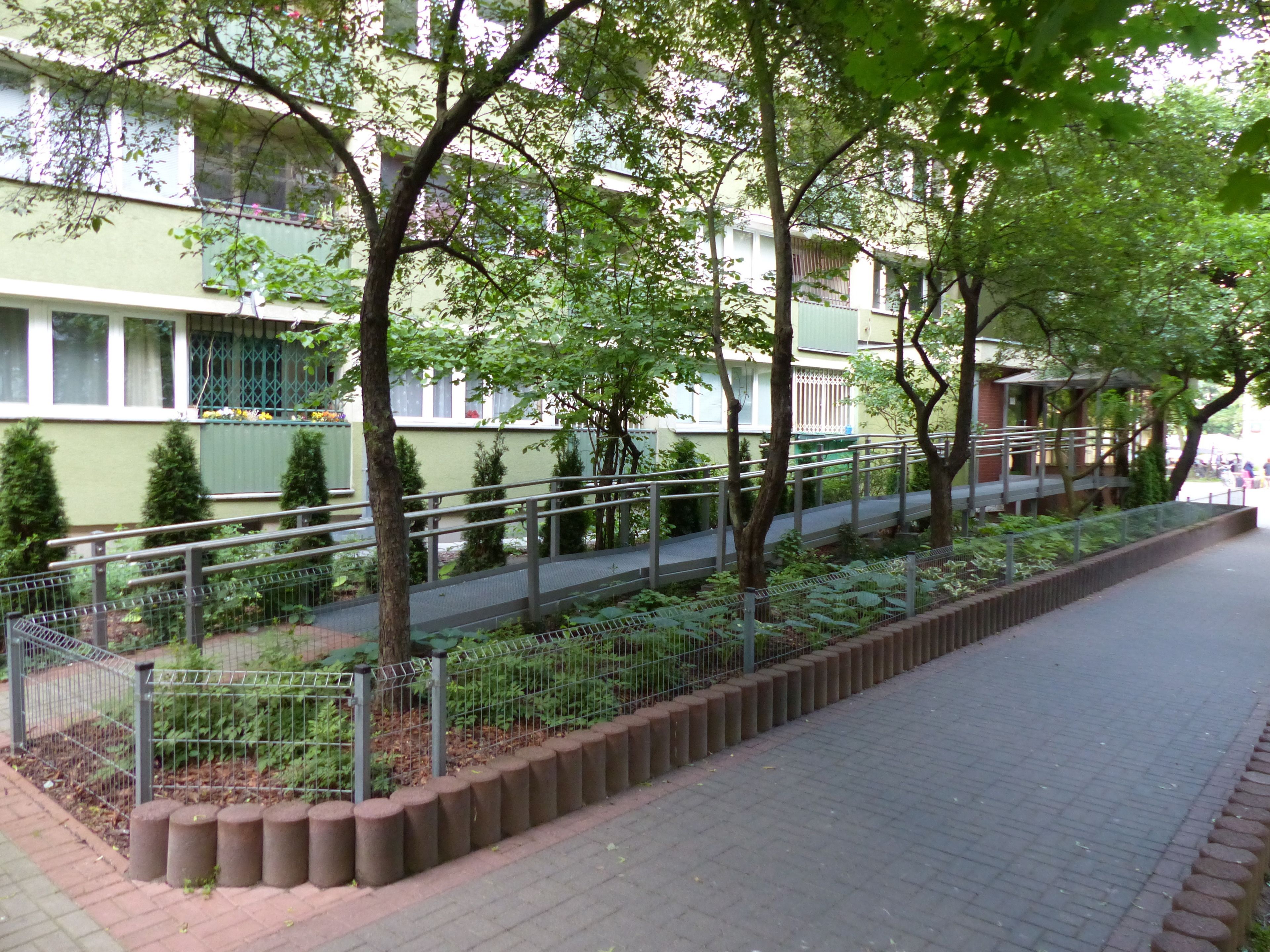 Un camino bordeado por árboles y una cerca de metal que conduce a un edificio con fachada verde y ventanas con contraventanas verdes.