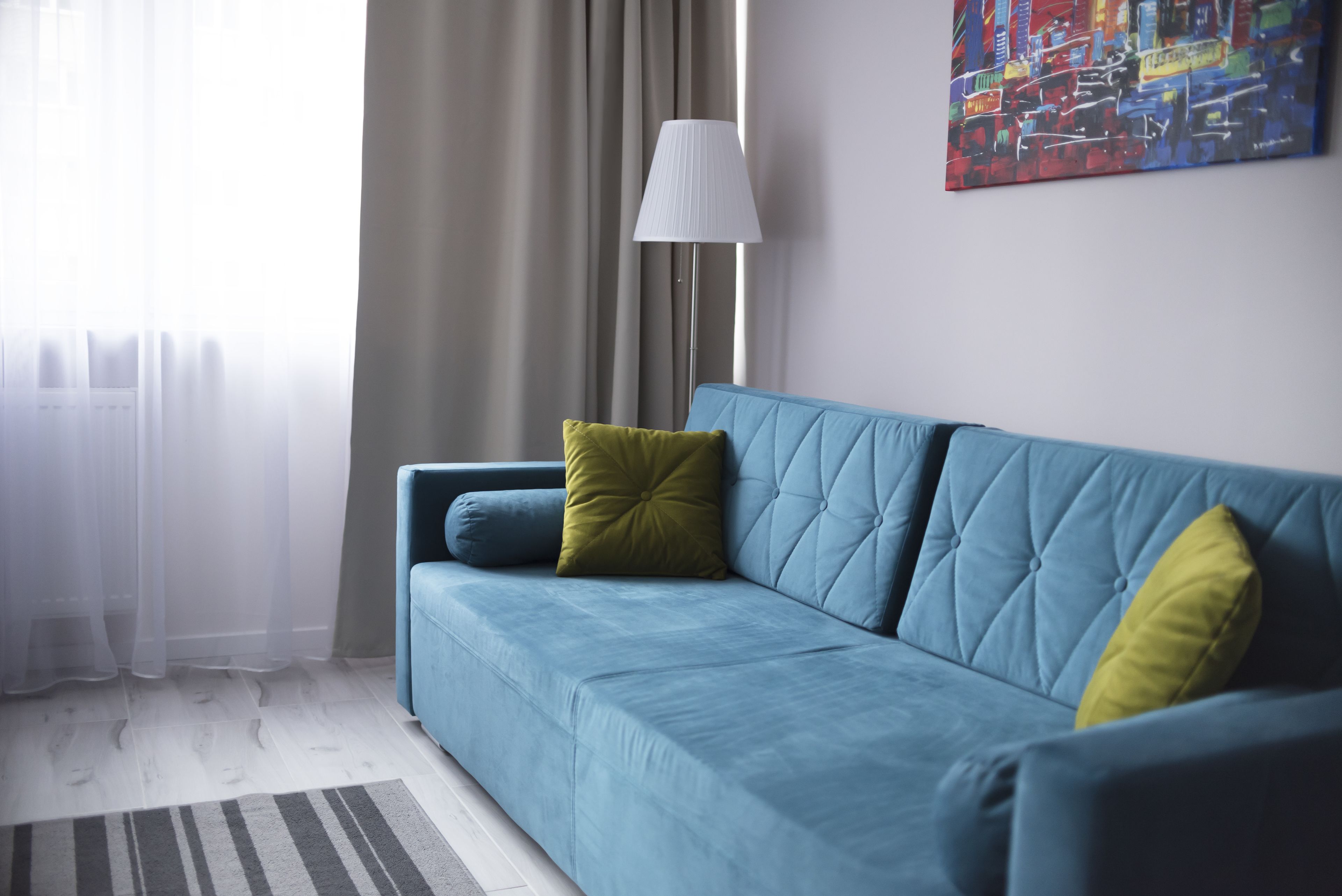 A modern living room with a blue sofa, a green pillow, a white lamp, and a colorful abstract painting on the wall.