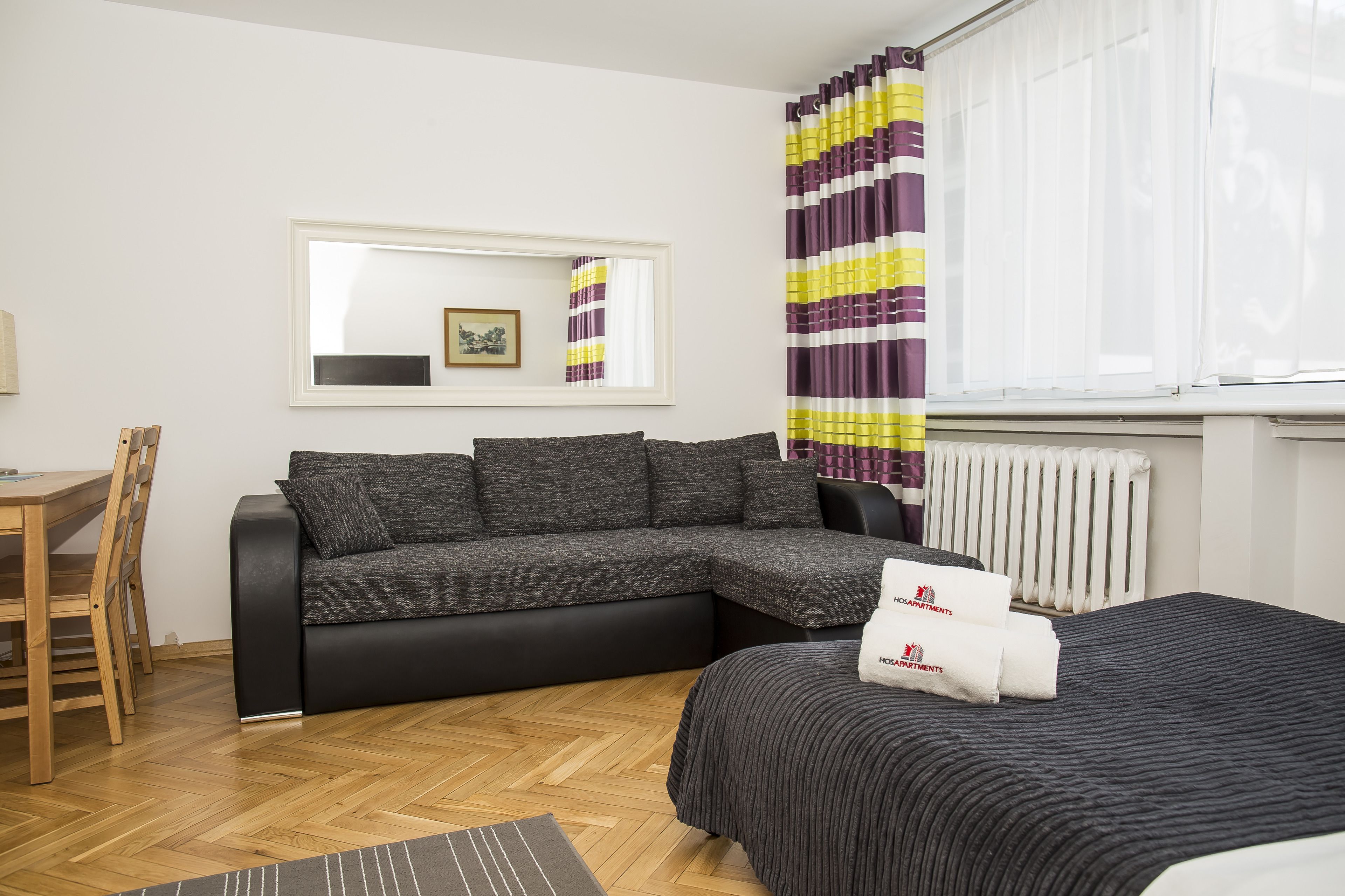A modern hotel room with a sofa, a bed, a wooden floor, a dining table, and a window with curtains.