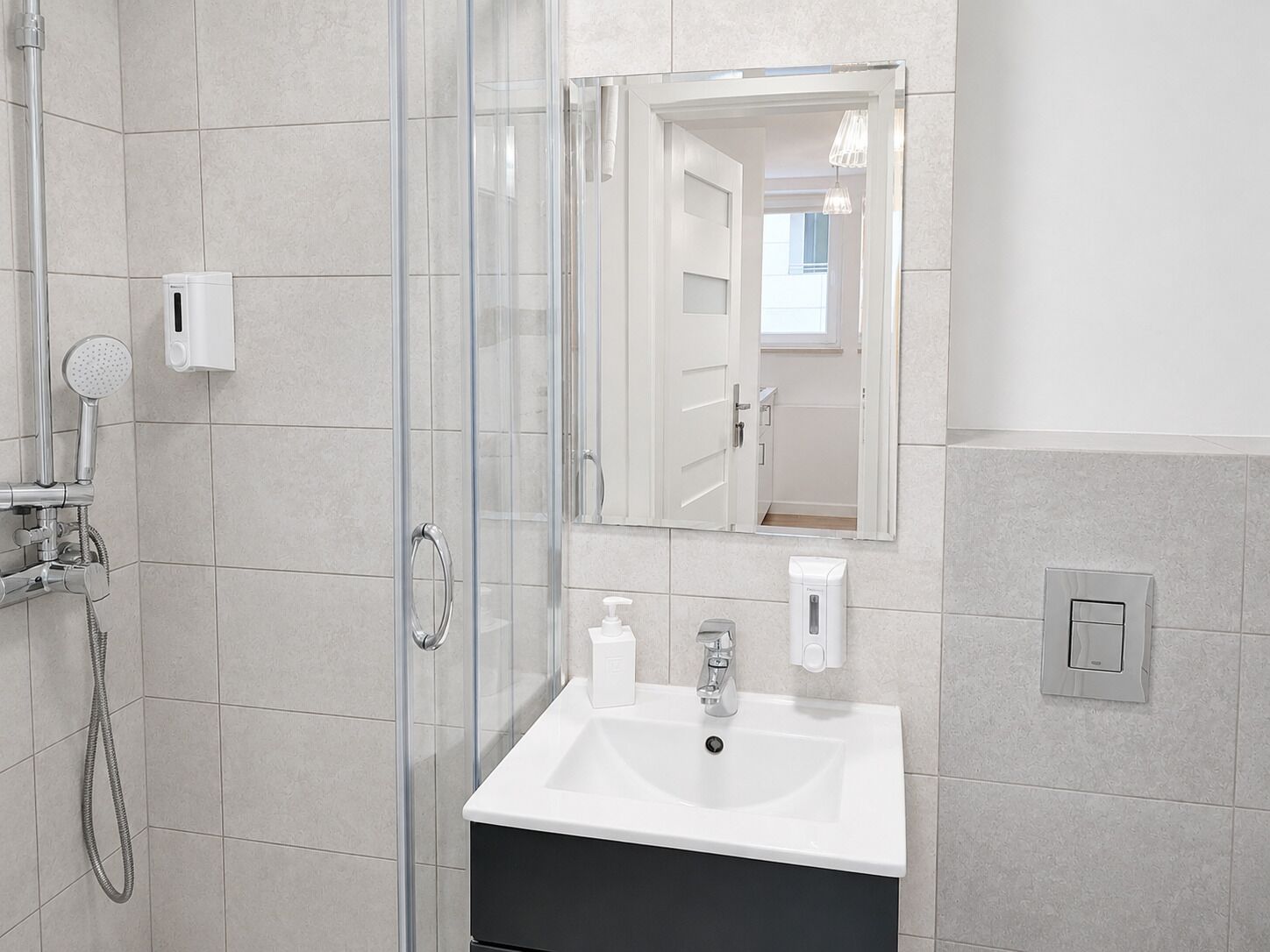 A modern bathroom with a glass shower enclosure, a white sink, and a mirror reflecting a door and a window.