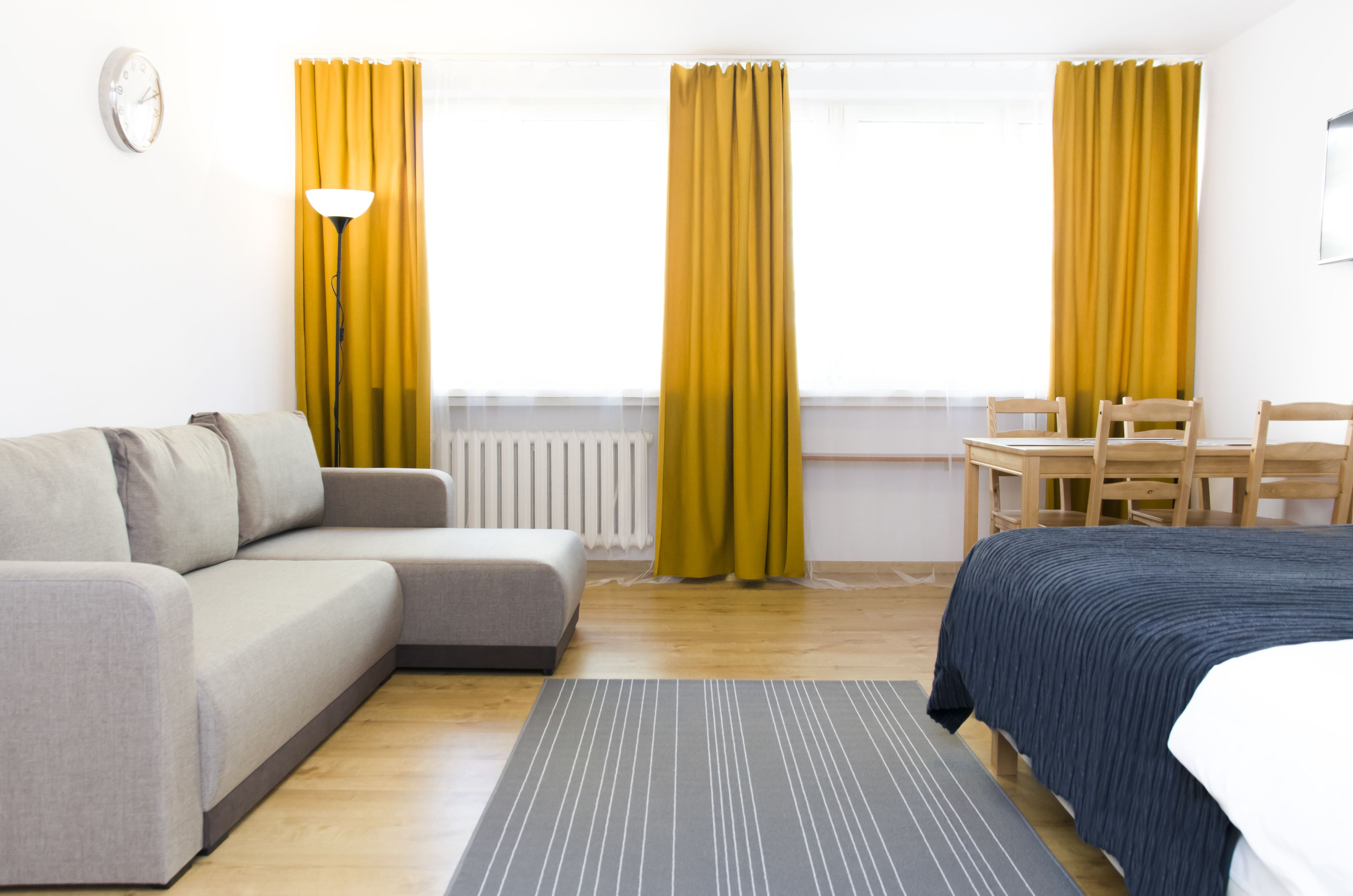 Ein modernes Hotelzimmer mit grauem Sofa, einem Bett mit dunkelblauer Bettdecke, einem Holztisch mit zwei Stühlen und gelben Vorhängen.