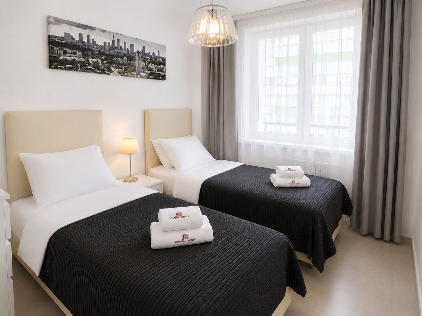A hotel room with two beds, a cityscape painting, a chandelier, and a window with curtains.
