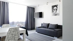 A modern living room with a grey sofa, a white dining table with geometric-patterned chairs, a flat-screen TV mounted on the wall, and a large window with grey curtains.