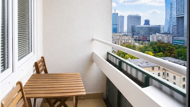 A balcony with a wooden table and chairs, a window with blinds, and a cityscape view.
