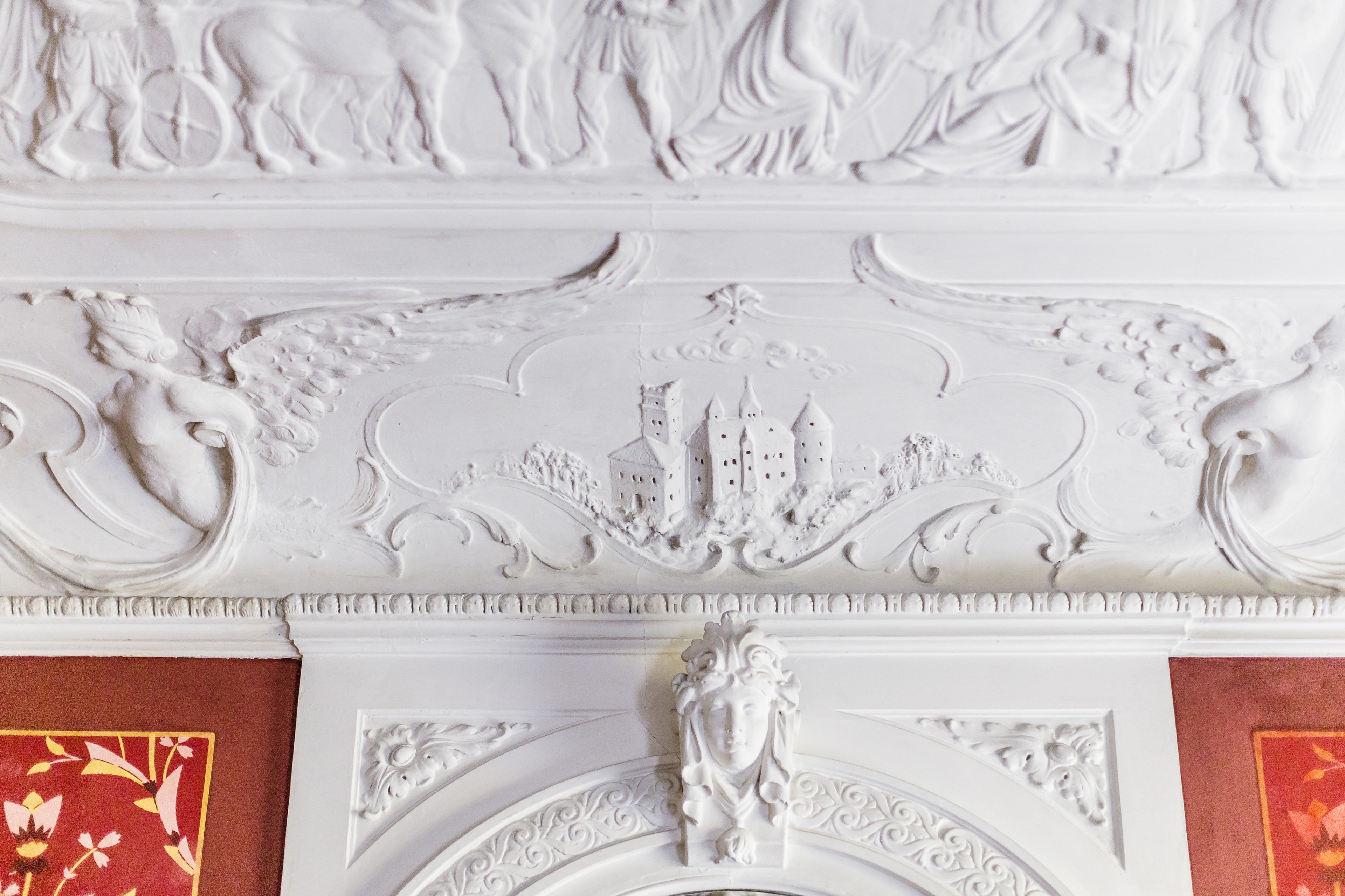 Ein detailliertes, weißes architektonisches Relief mit einer zentralen Figur und umlaufenden dekorativen Elementen.