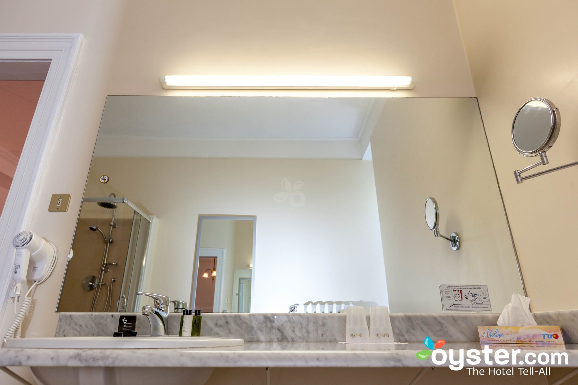 A bathroom with a large mirror, a sink, and a shower. There are toiletries on the counter and a towel rack on the wall.