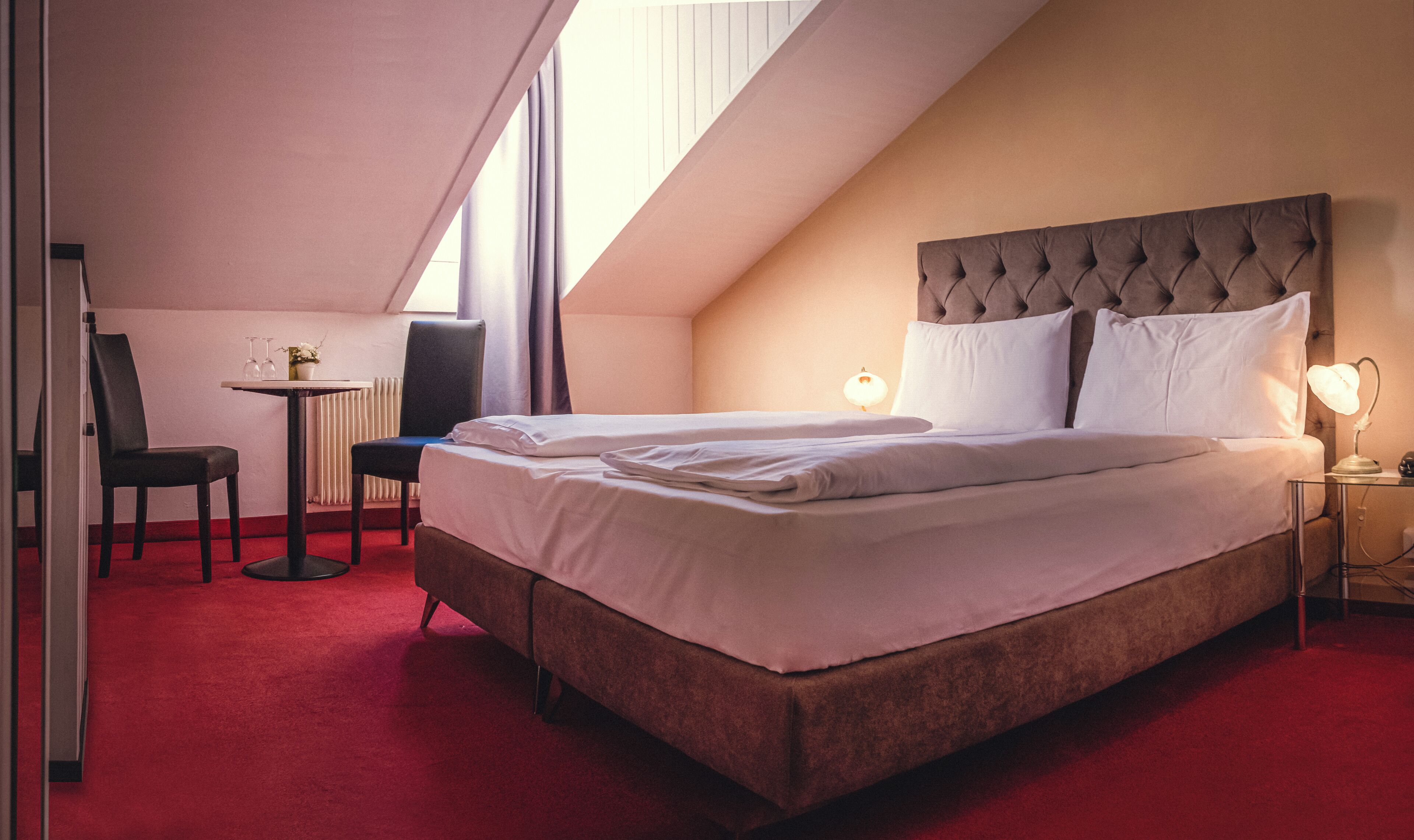 A hotel room with a large bed, a desk with a chair, and a small table with wine glasses.