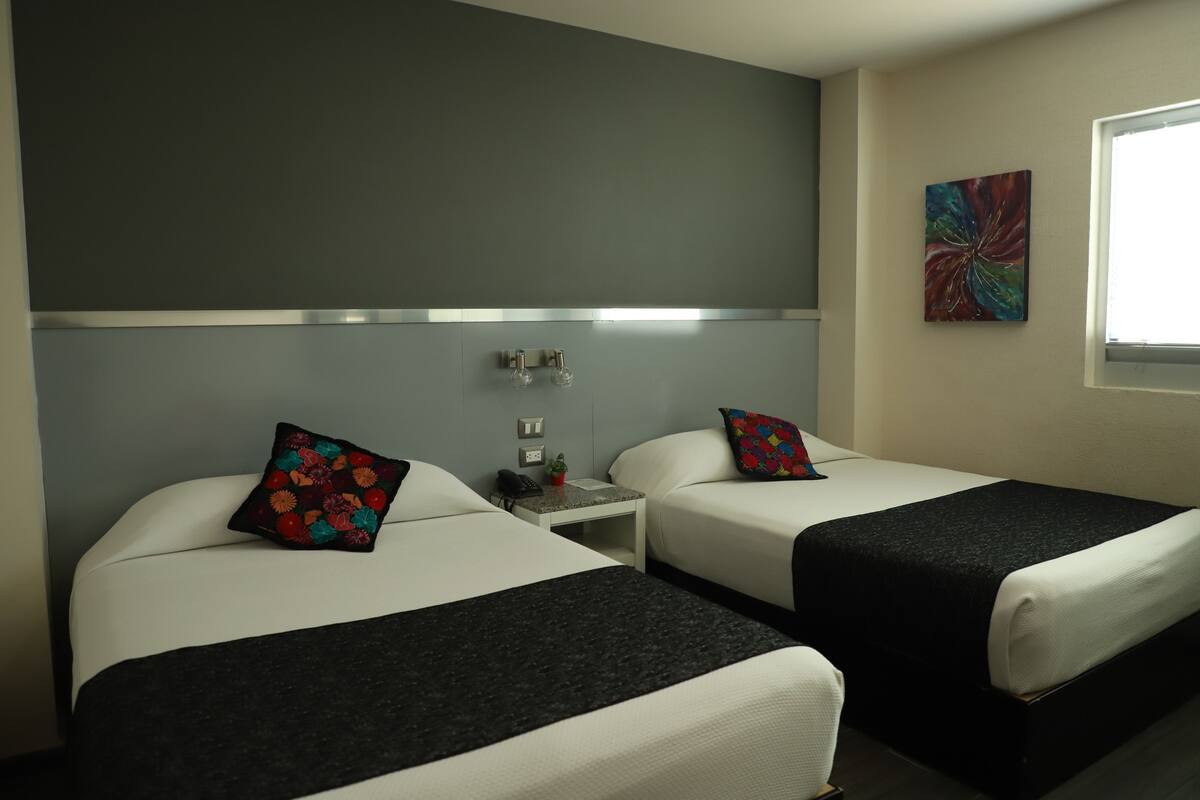 A hotel room with two beds, a nightstand, a phone, a small plant, and a window.