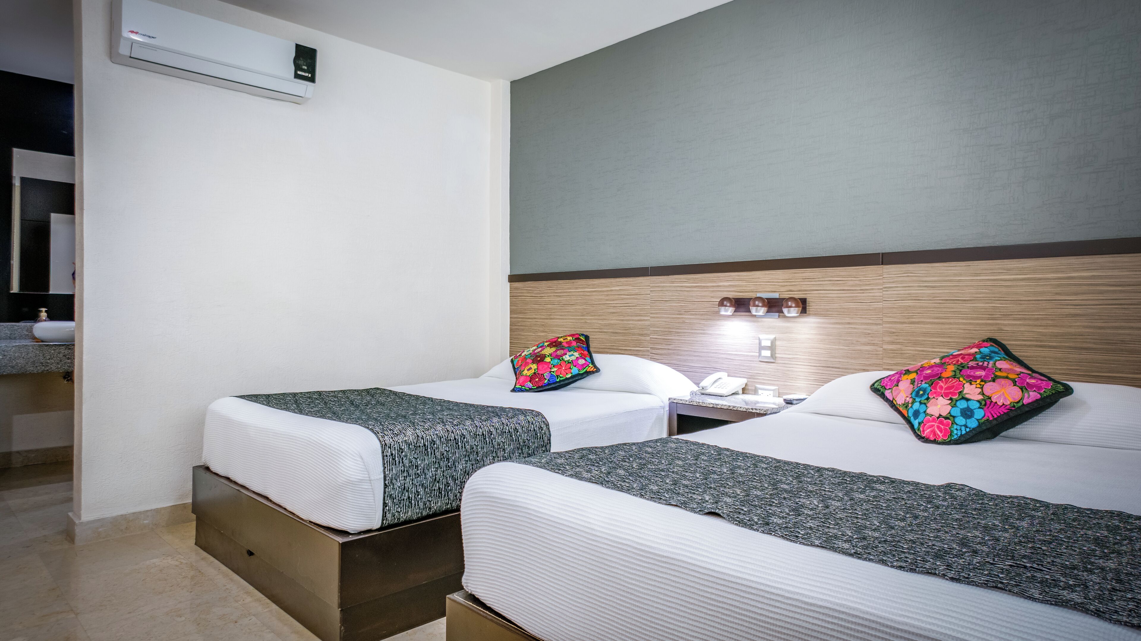 Habitación de hotel con dos camas, cada una con un cojín floral y una manta texturizada. Hay una mesita de noche con un teléfono y una lámpara. La habitación tiene un cabecero de madera y un aire acondicionado instalado en la pared.