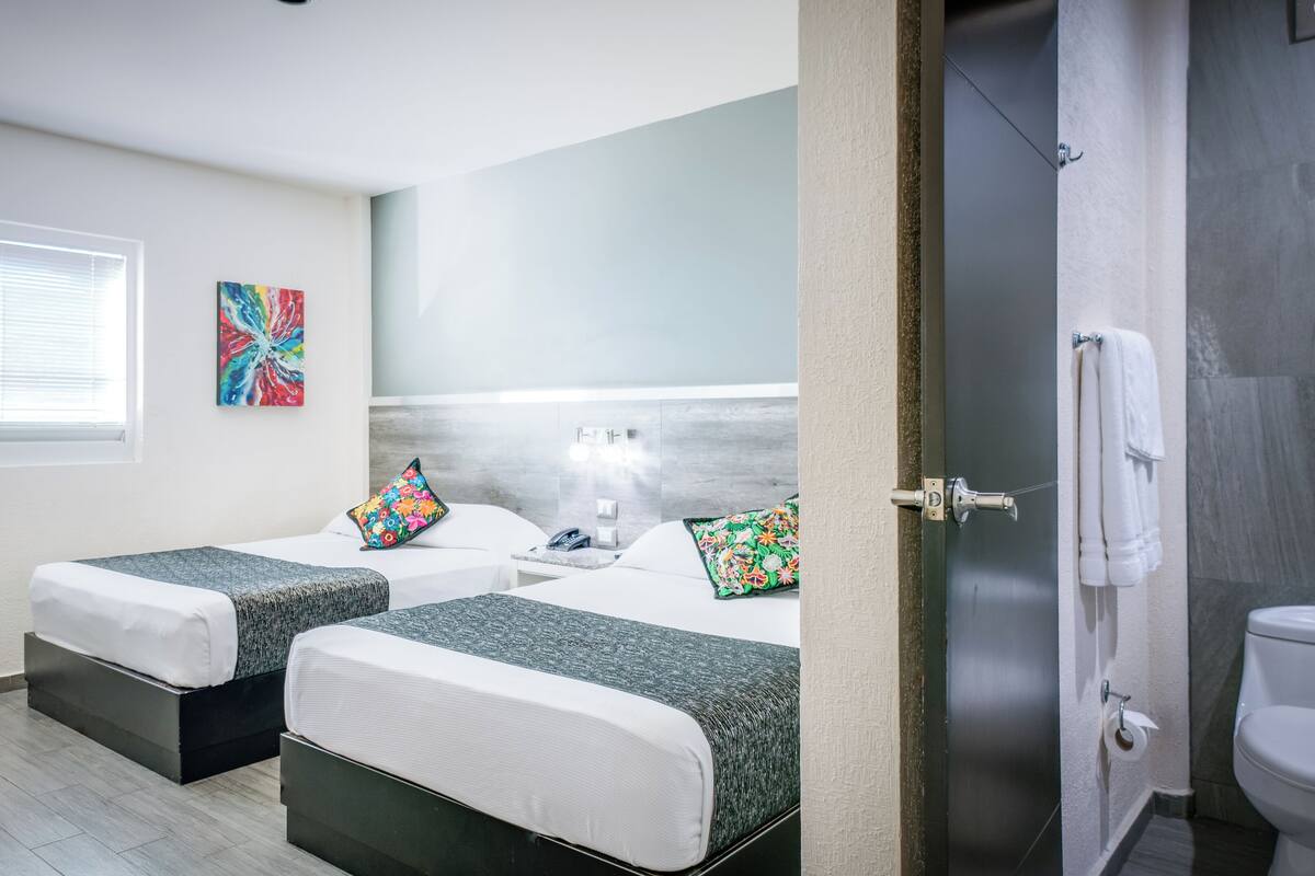 A hotel room with two beds, a window with blinds, a colorful abstract painting, and a bathroom visible through an open door.
