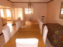 Una larga mesa de comedor con sillas blancas, un mantel estampado y una ventana con cortinas.