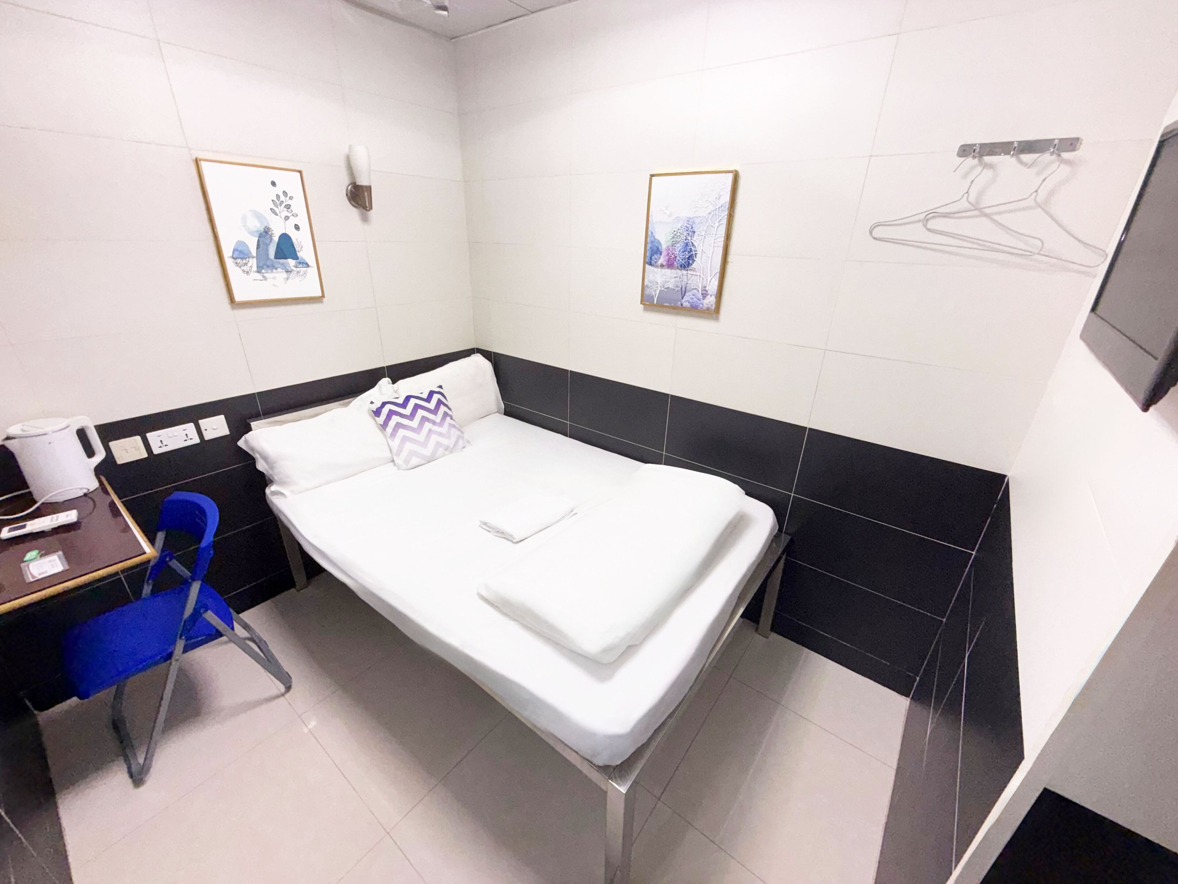 Ein kleines, sauberes Zimmer mit einem Bett, einem Schreibtisch und einem Stuhl. An der Wand hängen zwei gerahmte Bilder und ein Kleiderständer.