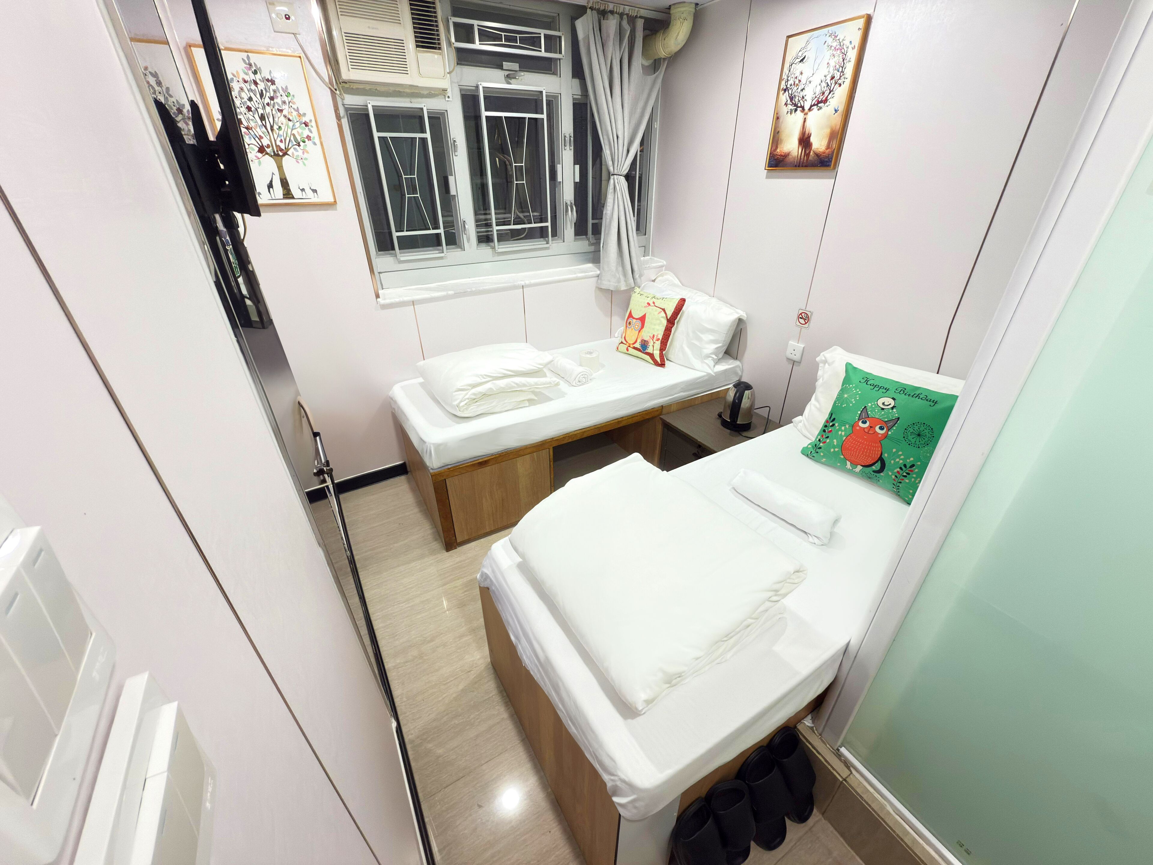 A small, clean room with two beds, a window with curtains, a radiator, and a green wall decoration.